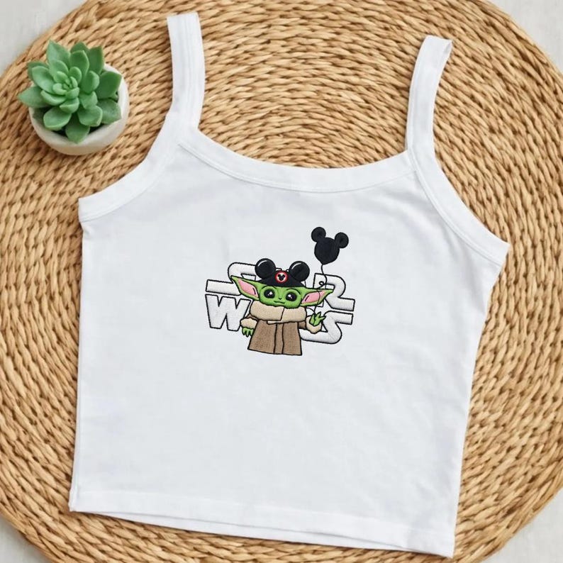 May include: White tank top with a graphic of Baby Yoda wearing Mickey Mouse ears and holding a black balloon. The Star Wars logo is behind the character. A small succulent plant in a white pot sits to the left.