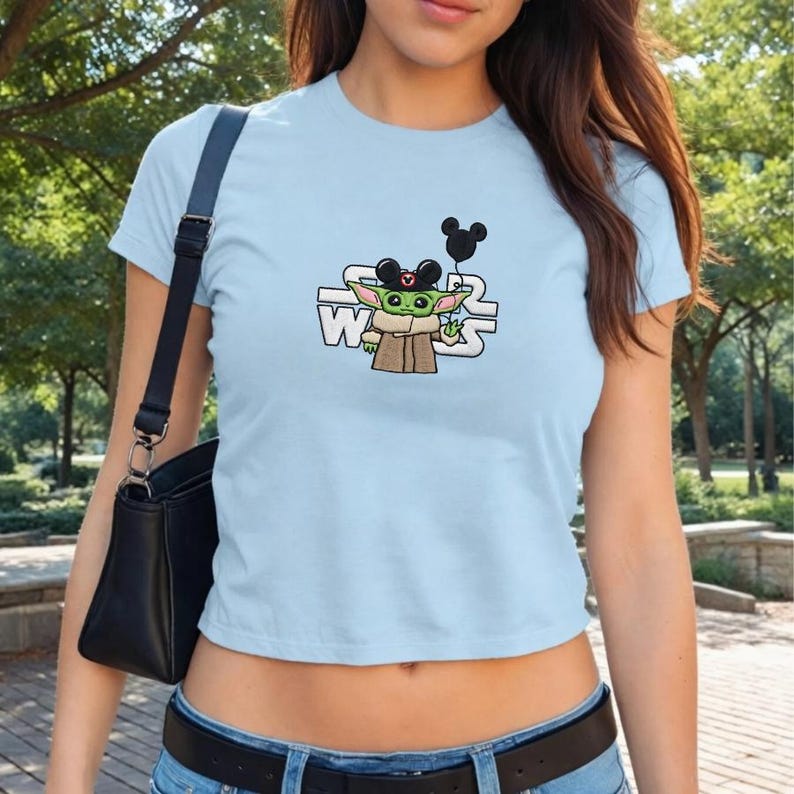 May include: Light blue crop top featuring a graphic of Grogu from Star Wars wearing Mickey Mouse ears and holding a black balloon. The Star Wars logo is printed in white. The shirt is a crew neck.
