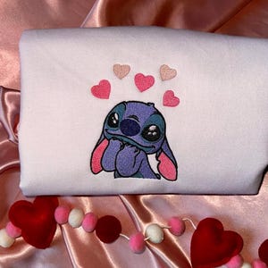 Puede incluir: Una sudadera blanca con una imagen bordada de Stitch de Lilo & Stitch, rodeada de corazones rosas. Una guirnalda de bolas y corazones de fieltro rojo, rosa y blanco decora el fondo.