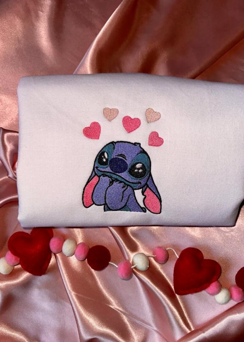 Puede incluir: Una sudadera blanca con una imagen bordada de Stitch de Lilo & Stitch, rodeada de corazones rosas. Una guirnalda de bolas y corazones de fieltro rojo, rosa y blanco decora el fondo.