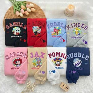 May include: A selection of folded sweatshirts in various colours, each with embroidered designs and text. The sweatshirts display the words "Gangle", "Caine", "Zooble", "Kinger", "Ragatha", "Jax", "Pomni", and "Bubble" alongside unique graphics. A neat arrangement.