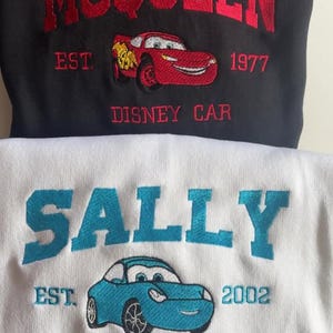 May include: Black sweatshirt with red embroidered text "MCQUEEN" above a red cartoon car and the text "DISNEY CAR". Below is a white sweatshirt with blue embroidered text "SALLY" above a blue cartoon car and the text "DISNEY CAR".