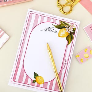 May include: A rectangular notepad with pink and white vertical stripes, featuring a central white oval area for writing labelled Notes. The design includes watercolour lemon illustrations with green leaves and white blossoms. A gold pen rests on the pad.