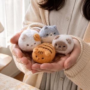 Pode incluir: Quatro pequenos brinquedos de pelúcia em forma de gato com texturas macias e felpudas. Um é branco com manchas laranja, um é azul claro, um é laranja com listras marrom-escuras e um é branco com rosto e orelhas marrons. Têm detalhes bordados.