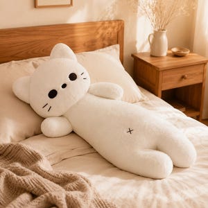 May include: A long, white plush cat pillow with a soft, fuzzy texture. The cat features two small, rounded ears, two black circular eyes, a small black circular nose, and three black embroidered whiskers on each side. A small black X is stitched on the body.
