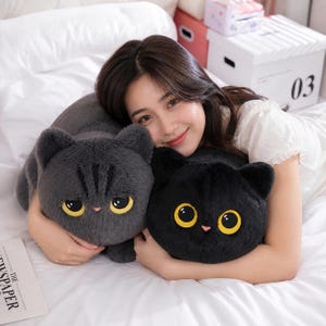 Pode incluir: Duas almofadas de pelúcia em forma de gato. Uma é cinza escuro, a outra preta. Ambas têm olhos grandes amarelos e pequenos narizes rosa. As almofadas estão em uma colcha branca.