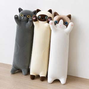 May include: Three long, cylindrical plush cat pillows standing upright. The left cat is dark gray with yellow eyes, the middle cat is cream and brown with blue eyes, and the right cat is white and brown with blue eyes. Each has raised paws.