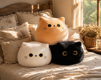 May include: Three plush cat-shaped cushions in white, tan, and black. Each cushion features a rounded head, pointed ears, large circular yellow eyes with black pupils, a small black nose, and three stitched whiskers on each side of the face.