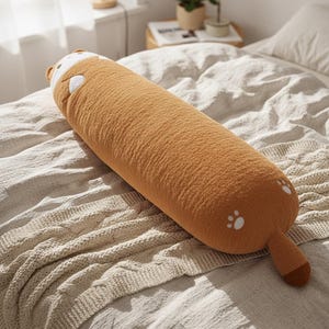May include: A long, brown plush dog-shaped pillow. It features a white face, ears, and paw prints on the end. The pillow is placed on a bed with a cream-coloured knitted blanket.