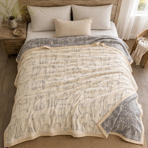 May include: A cream-coloured cotton quilt featuring a repeating pattern of grey line-drawn cats. The quilt has a wide grey border with a matching cat print and is finished with a solid cream-coloured fabric trim along the edges of the blanket.