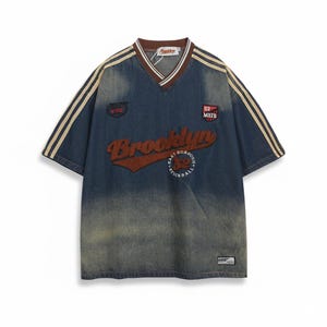 May include: A blue denim-style jersey with a V-neck and short sleeves. The jersey features the word "Brooklyn" in a brown, textured font across the chest, with additional text and patches. Beige stripes run down the sleeves.