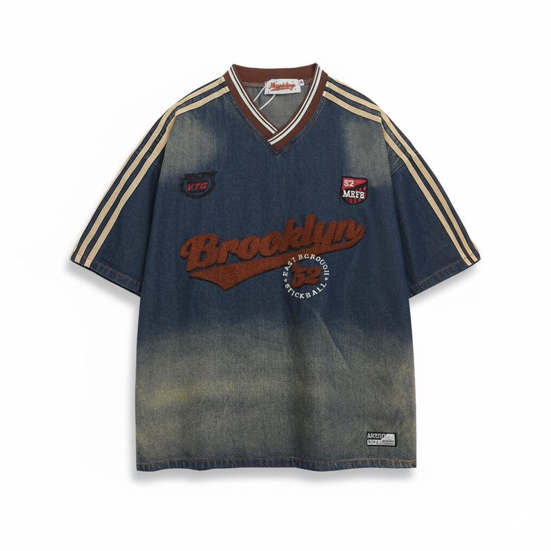 May include: A blue denim-style jersey with a V-neck and short sleeves. The jersey features the word "Brooklyn" in a brown, textured font across the chest, with additional text and patches. Beige stripes run down the sleeves.