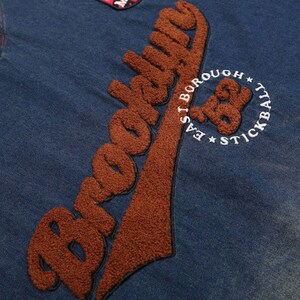 May include: Close-up of a blue denim jacket with brown embroidered text that says "Brooklyn". The jacket also features a patch with the text "East Borough Stickball".