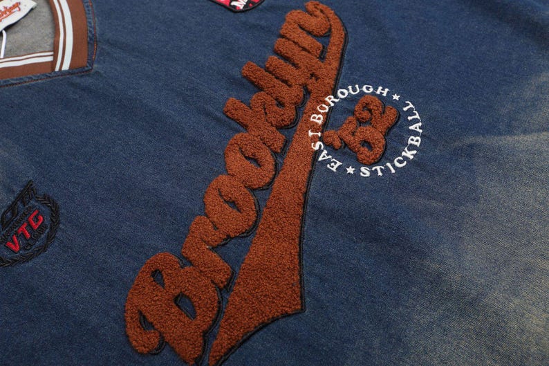 May include: Close-up of a blue denim jacket with brown embroidered text that says "Brooklyn". The jacket also features a patch with the text "East Borough Stickball".