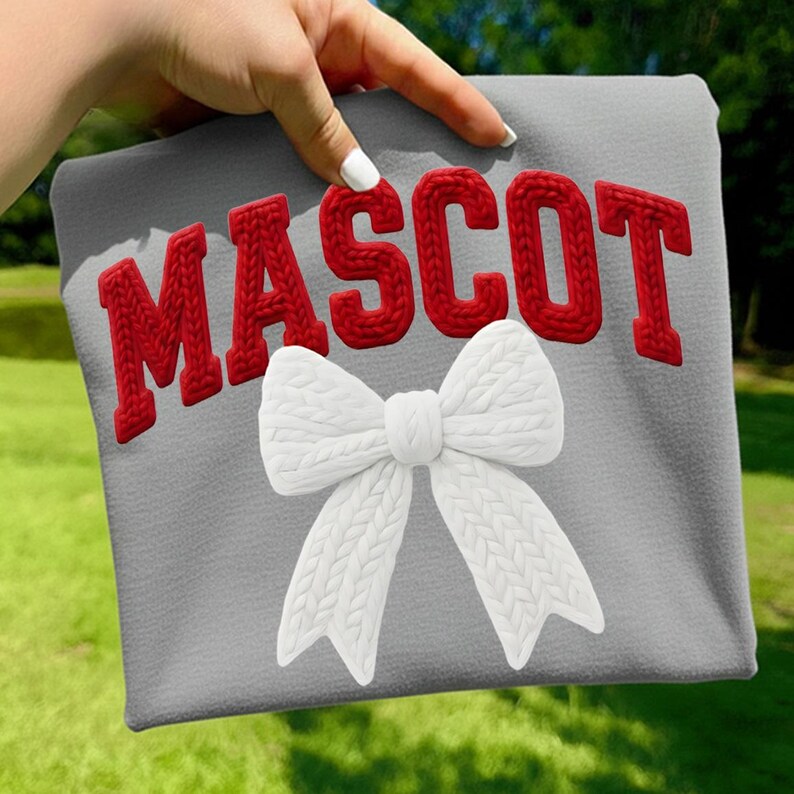 May include: A gray sweatshirt with the word "MASCOT" in red, textured letters. A white, knitted bow is centered below the text. The sweatshirt is held up against a green, grassy background.