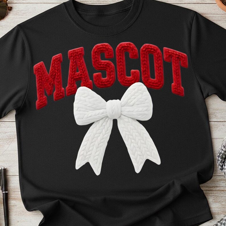 May include: Black t-shirt with the word "MASCOT" in red, textured, knitted letters. A large, white, knitted bow is centered below the text. The shirt is a casual style.