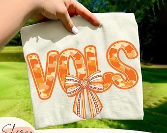 Może przedstawiać: Kremowa koszulka z napisem "VOLS" w pomarańczowych literach moro i pasującą grafiką kokardy. Tekst "PNG DIGITAL DOWNLOAD" znajduje się pod koszulką. Projekt jest trzymany przez dłoń.