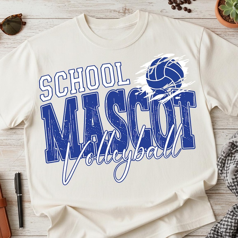 K&ouml;nnte beinhalten: Cremefarbenes T-Shirt mit den Worten "SCHOOL MASCOT Volleyball" in Blau und Wei&szlig;. Eine blaue Volleyball-Grafik befindet sich auf der rechten Seite. Das Shirt ist aus einem weichen Material.
