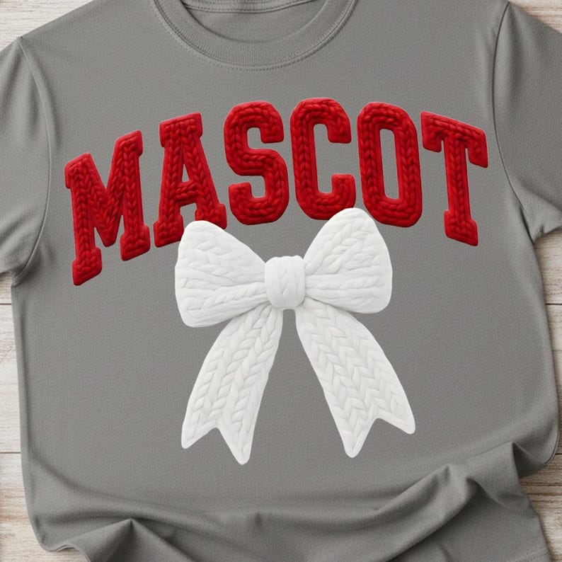 May include: Gray t-shirt with the word "MASCOT" in red, textured, block letters across the top. A white, knitted bow is centered below the word. The shirt is made of a soft material.