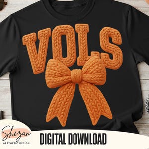 May include: Black t-shirt featuring the word "VOLS" in orange, textured, knitted-style letters above an orange bow. The words "DIGITAL DOWNLOAD" are printed in white on a black background. The design is by Shezan Aesthetic Design.