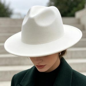 Puede incluir: Un sombrero fedora de color blanco sólido con una corona estructurada y un ala ancha y plana. El sombrero se combina con un abrigo de mezcla de lana verde oscuro con solapas de muesca y una prenda superior negra de cuello alto.