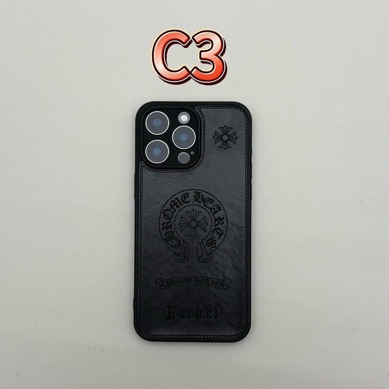 Pu&ograve; includere: Custodia per telefono in pelle nera con logo Chrome Hearts in rilievo e design floreale. La custodia ha un ritaglio per la fotocamera con tre obiettivi e un flash. La parola "Roobel" &egrave; stampata in basso. Un logo "C3" rosso e arancione &egrave; in alto.