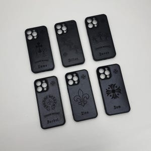 May include: A collection of six black phone cases with various embossed designs, including crosses, fleur-de-lis, and a heart emblem. Each case features a different name: Emma, Dillon, Susan, Rachel, Elsa, and Sam.