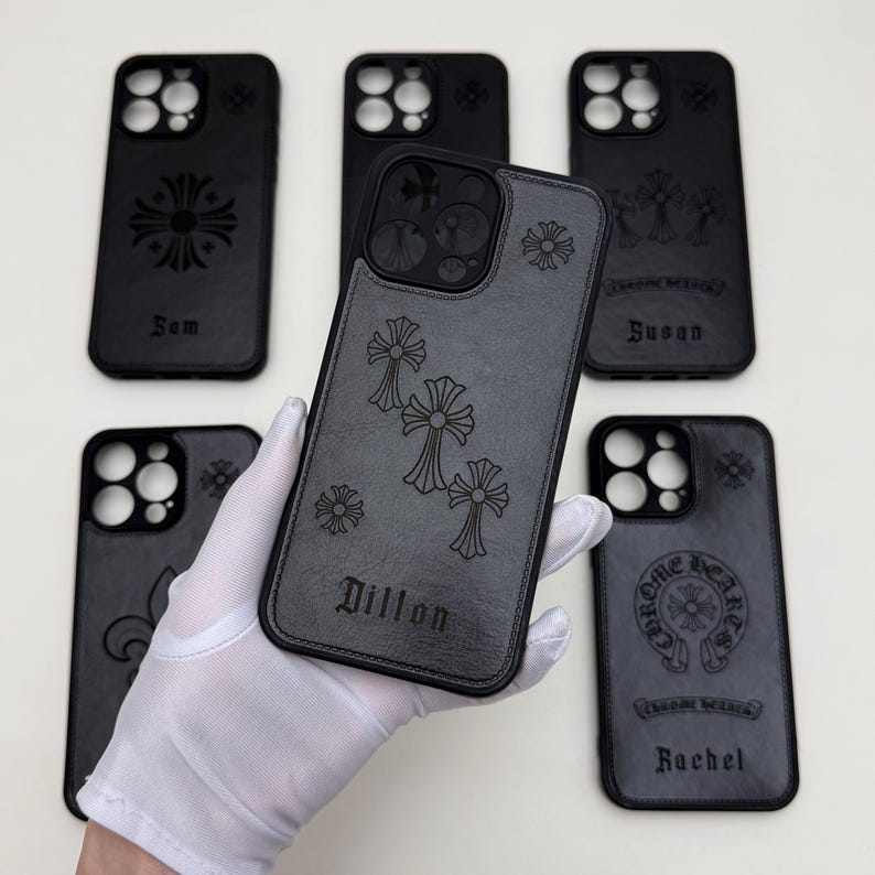 May include: A collection of black phone cases with engraved cross and floral designs. Some cases feature names like "Sam", "Dillon", "Susan", and "Rachel". The cases are made of a dark gray material.