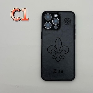 May include: Black phone case with a fleur-de-lis design and the name "Elsa" embossed on the lower part. The case has a black border and a camera cutout. A small floral design is on the upper right corner. The text "C1" is in the upper left corner.