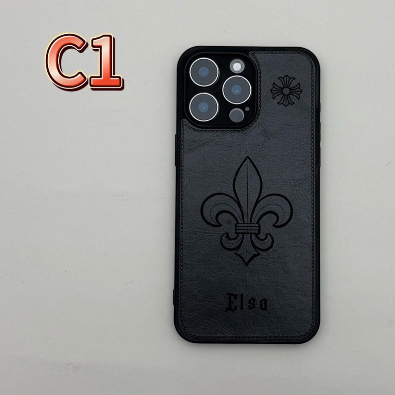 May include: Black phone case with a fleur-de-lis design and the name "Elsa" embossed on the lower part. The case has a black border and a camera cutout. A small floral design is on the upper right corner. The text "C1" is in the upper left corner.