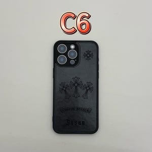 Pu&ograve; includere: Custodia per telefono in pelle nera con disegni a croce in rilievo e la scritta "CHROME HEARTS" e "Susan". La custodia ha un ritaglio per la fotocamera e una croce decorativa in alto. Il testo "C6" &egrave; sopra.