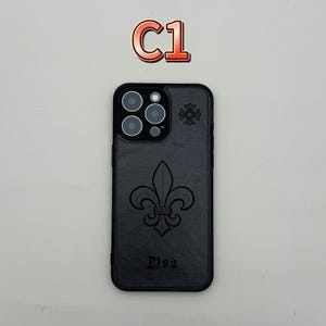 Pu&ograve; includere: Custodia per telefono nera con un design a giglio e il nome "Elsa" in rilievo in basso. La custodia presenta un motivo floreale vicino alla fotocamera e il testo "C1" in alto.
