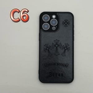 May include: Black phone case with embossed cross designs and the text "CHROME HEARTS" and "Susan". The case has a black camera lens surround and a stitched leather-like texture. The upper left corner has the text "C6".