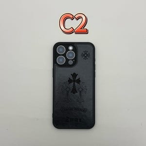 Pu&ograve; includere: Custodia per telefono in pelle nera con un design a croce decorativo e il nome "Emma" in rilievo in basso. La custodia ha un bordo nero e un ritaglio per l'obiettivo della fotocamera. Il testo "C2" &egrave; sopra.