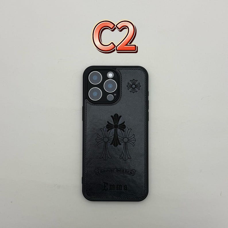 Pu&ograve; includere: Custodia per telefono in pelle nera con un design a croce decorativo e il nome "Emma" in rilievo in basso. La custodia ha un bordo nero e un ritaglio per l'obiettivo della fotocamera. Il testo "C2" &egrave; sopra.