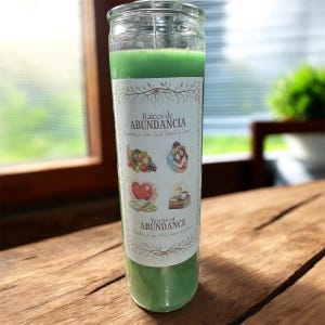 May include: A green candle in a clear glass jar with a label that reads "Raices de Abundancia" and "Roots of Abundance." The label features illustrations of a cornucopia, a couple, a heart, and books. The candle sits on a wooden surface.