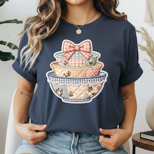 May include: Navy blue t-shirt featuring a stack of decorative bowls with a bow on top. The bowls have a patchwork design with floral and polka dot patterns. The shirt is a casual style.