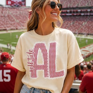 以下が含まれることがあります： クリーム色の半袖Tシャツで、「roll tide」という文字と、濃い赤と白の縦縞模様の「AL」という文字がプリントされています。文字は白いスカラップ状のレースのような縁取りで囲まれています。