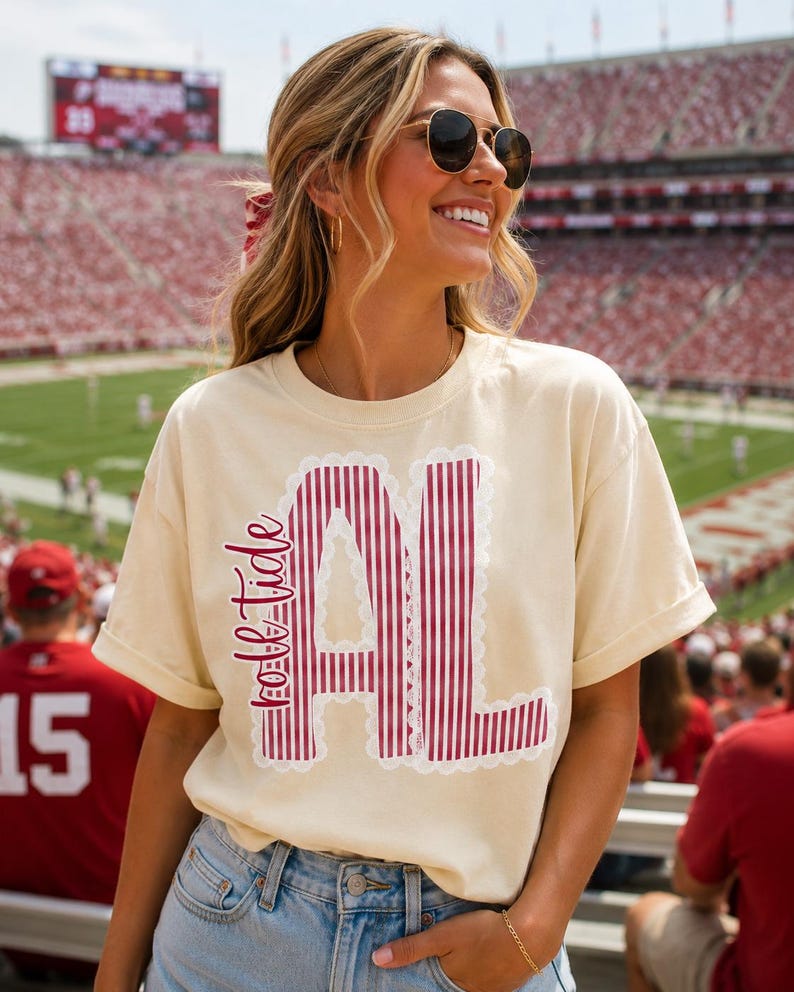 以下が含まれることがあります： クリーム色の半袖Tシャツで、「roll tide」という文字と、濃い赤と白の縦縞模様の「AL」という文字がプリントされています。文字は白いスカラップ状のレースのような縁取りで囲まれています。