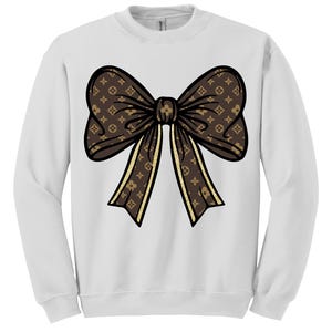 Puede incluir: Sudadera blanca con un gran dise&ntilde;o de lazo marr&oacute;n. El lazo tiene un patr&oacute;n repetitivo y detalles dorados. El lazo est&aacute; centrado en la parte delantera de la sudadera.
