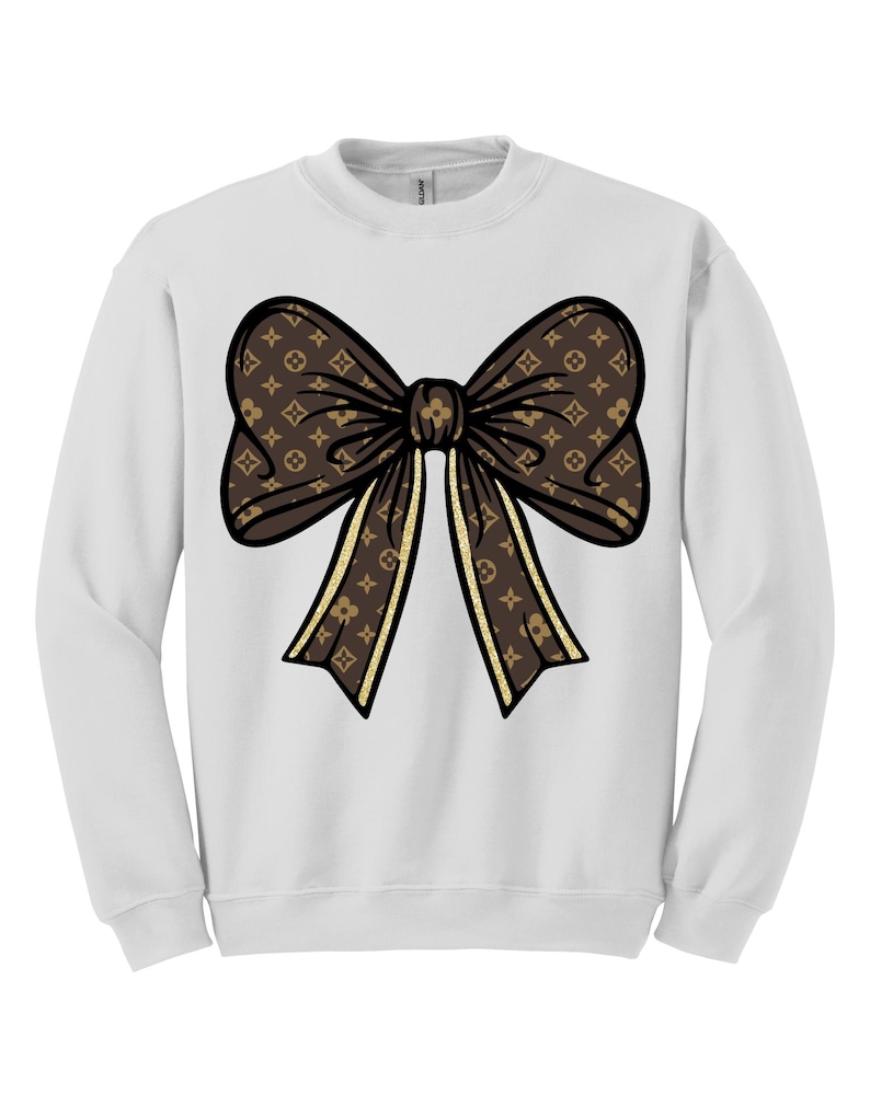 Puede incluir: Sudadera blanca con un gran dise&ntilde;o de lazo marr&oacute;n. El lazo tiene un patr&oacute;n repetitivo y detalles dorados. El lazo est&aacute; centrado en la parte delantera de la sudadera.