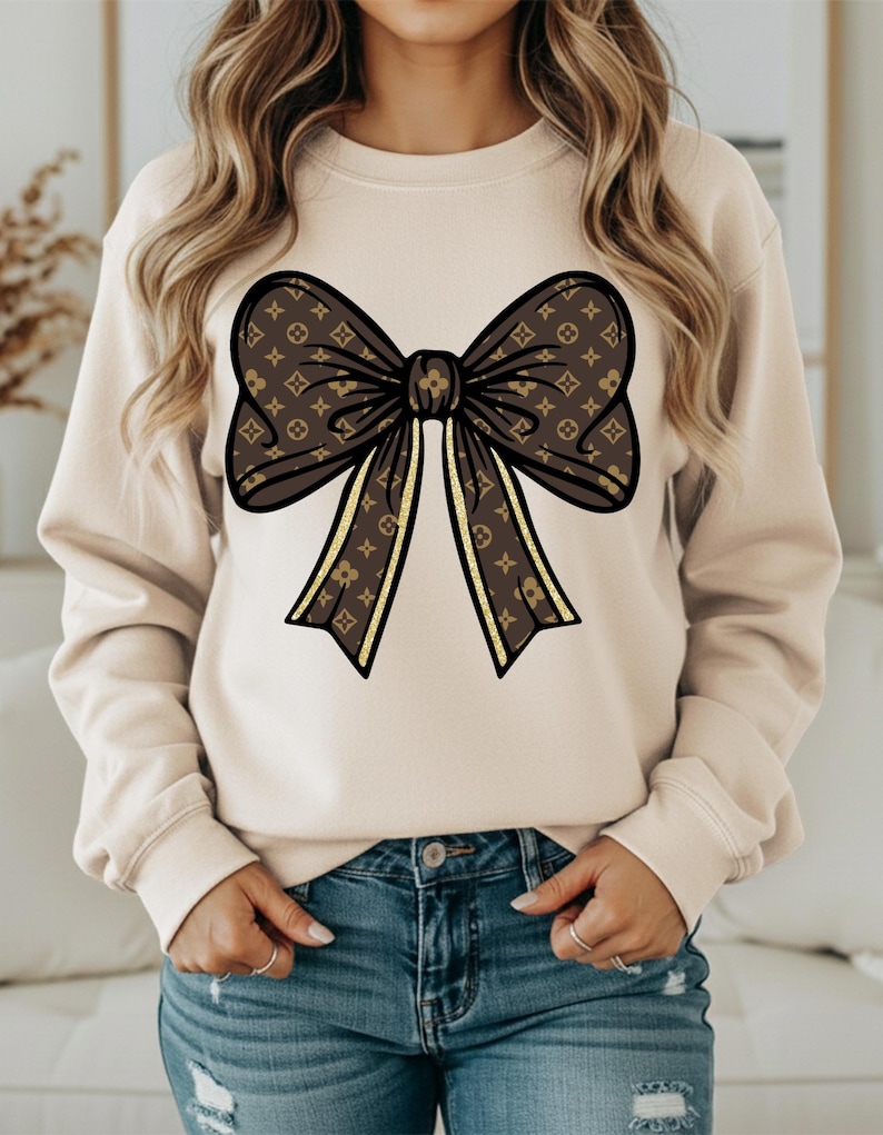 Puede incluir: Sudadera beige con un gran lazo marr&oacute;n con un patr&oacute;n repetido y detalles dorados. El lazo est&aacute; centrado en la parte delantera. La modelo lleva vaqueros azules.
