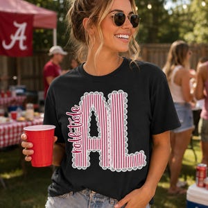以下が含まれることがあります： 「roll tide」というテキストと「AL」という文字がデザインされた黒の半袖Tシャツ。文字は赤と白の縦縞模様で塗りつぶされており、白いレースのような縁取りが施されています。