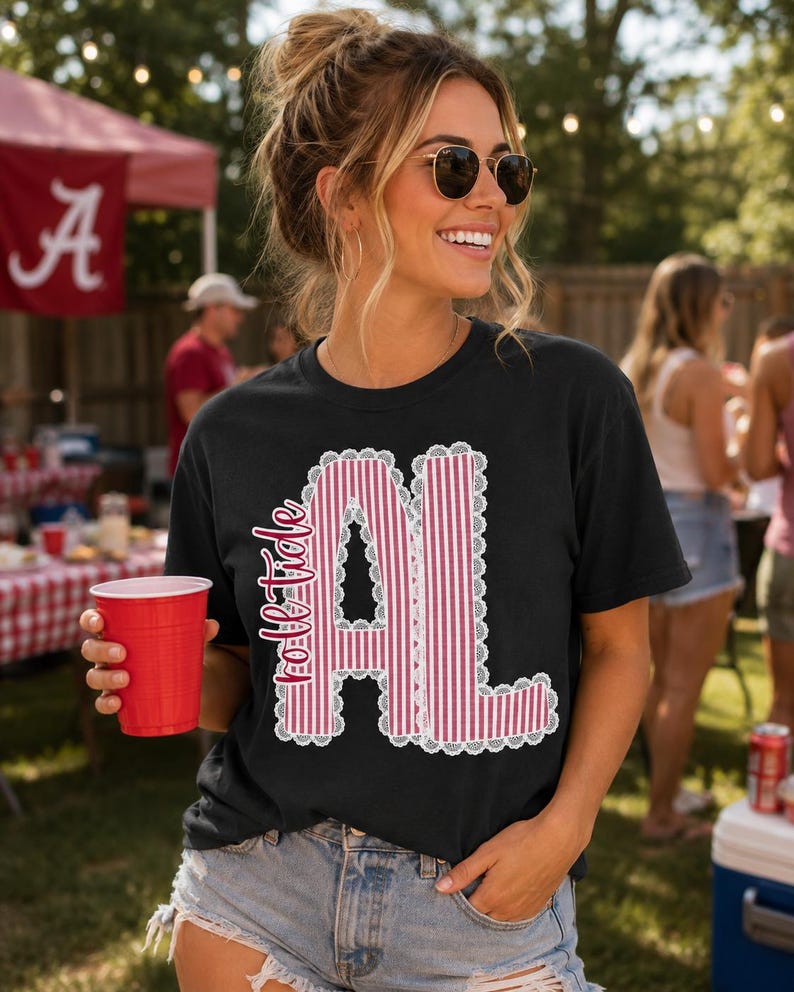以下が含まれることがあります： 「roll tide」というテキストと「AL」という文字がデザインされた黒の半袖Tシャツ。文字は赤と白の縦縞模様で塗りつぶされており、白いレースのような縁取りが施されています。