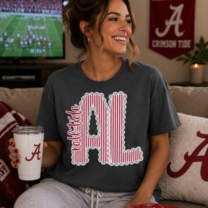 以下が含まれることがあります： チャコールグレーの半袖Tシャツ。前面には「Roll Tide」の文字と「AL」の文字が赤と白の縦縞模様で描かれ、白いレースのような縁取りが施されたグラフィックデザインがあしらわれています。