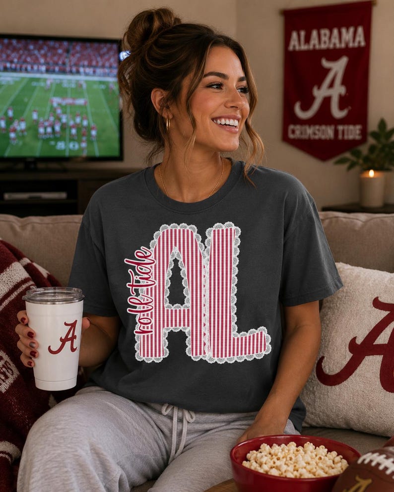以下が含まれることがあります： チャコールグレーの半袖Tシャツ。前面には「Roll Tide」の文字と「AL」の文字が赤と白の縦縞模様で描かれ、白いレースのような縁取りが施されたグラフィックデザインがあしらわれています。