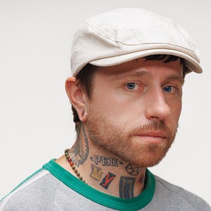 May include: A light beige newsboy cap with a curved brim. The person is wearing a gray and green shirt and has tattoos on their neck. The person has a beard and is looking at the camera.