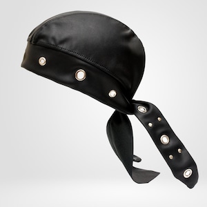 May include: A black durag-style head covering made of a smooth, semi-glossy material. The headpiece features a wide band adorned with evenly spaced silver-toned metal grommets and includes two long, trailing fabric ties that also feature grommets.