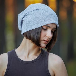May include: A light gray beanie hat. The hat is made of a soft, knitted material and is worn on the head. The person is wearing a dark gray tank top. The background is blurred with shades of green and brown.