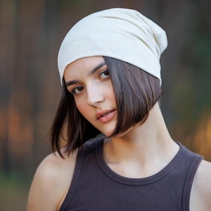 May include: A solid off-white beanie hat made of a soft, lightweight fabric. The beanie features a simple, pull-on design that fits snugly around the head, providing a clean and smooth appearance without any visible patterns, logos, or textures.